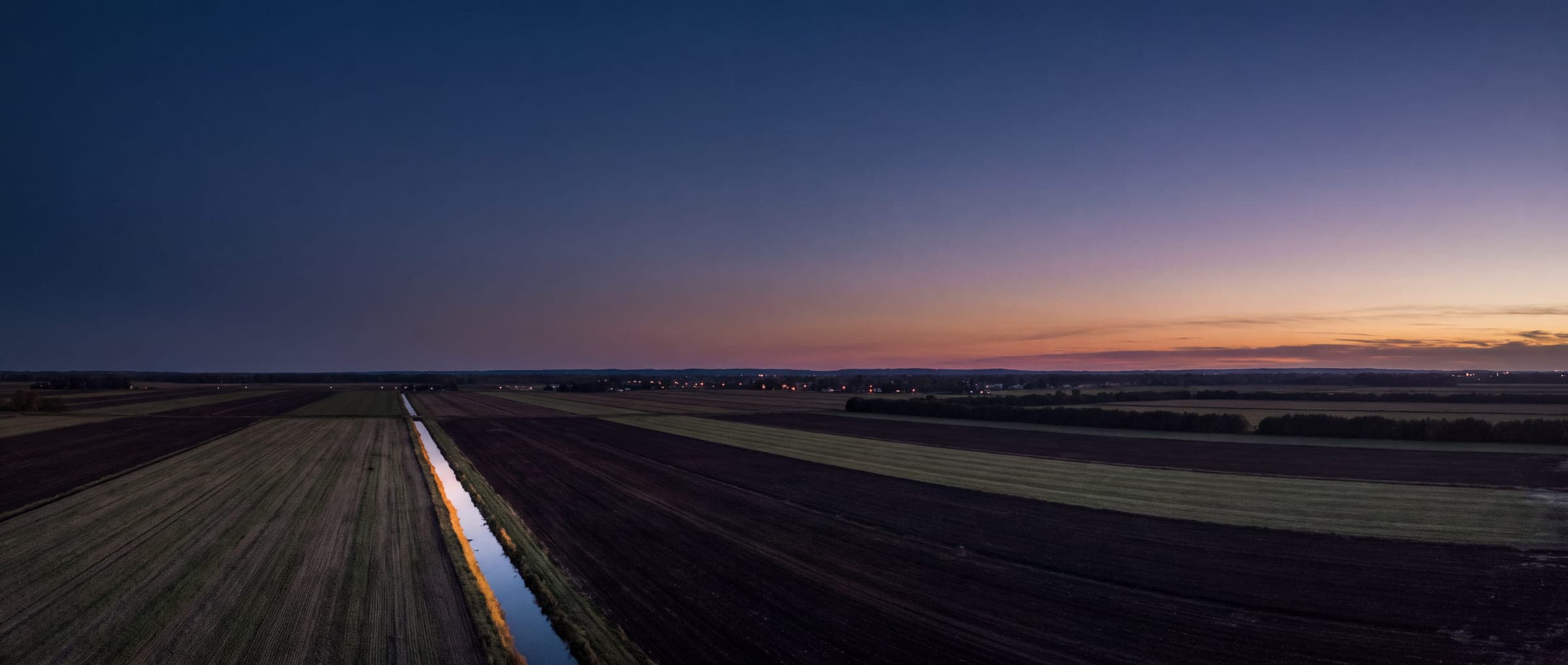 Aerial view of flat marsh farmland divided into strips, with a straight drainage canal catching the last light after sunset